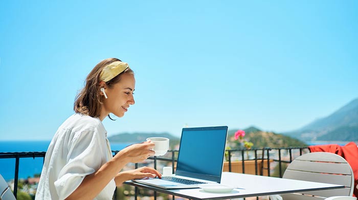Kvinna arbetar avslappnat med laptop och kaffe på ett café på en resort – frilansliv i solig miljö.