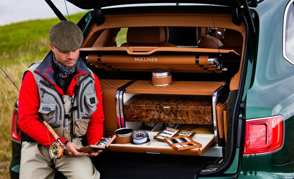 En man i fiskeväst sitter vid den öppna bakluckan på en grön Bentley Bentayga och plockar bland flugaskar. Bagageutrymmet är inrett med läderklädda spötuber, flugaskar och lådor i rotfanér.