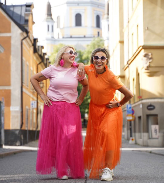 Lena Togay och Maria Grins skrattar tillsammans på en stadsgata, klädda i rosa och orange.