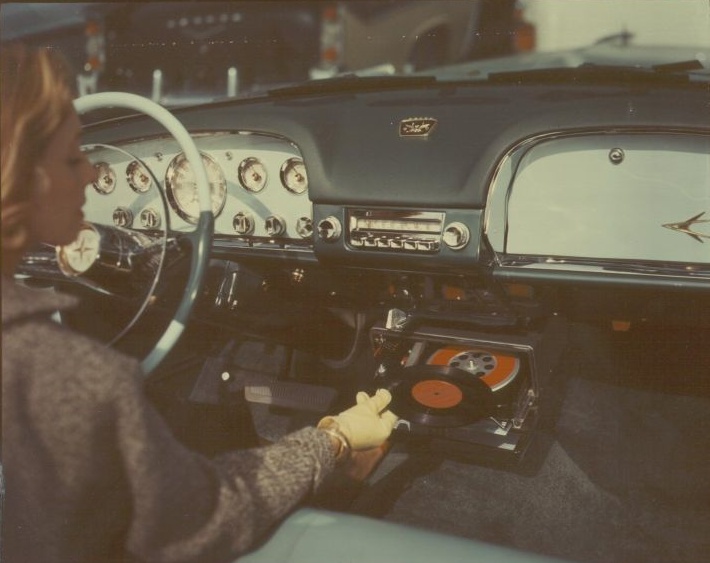 En kvinna i en DeSoto av 1956 års modell lägger en skiva i skivspelaren Highway Hi-Fi under instrumentbrädan.