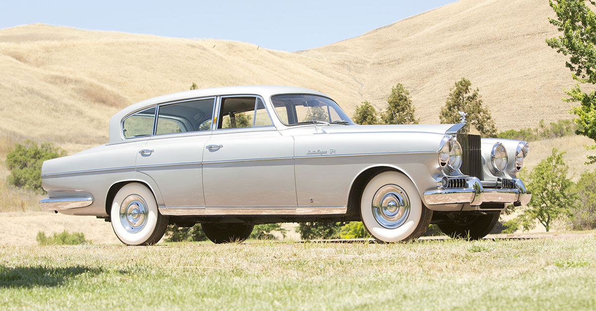 En silverfärgad Rolls-Royce Silver Wraith från 1954 med fyra strålkastare och vita däcksidor, parkerad framför gula kullar.